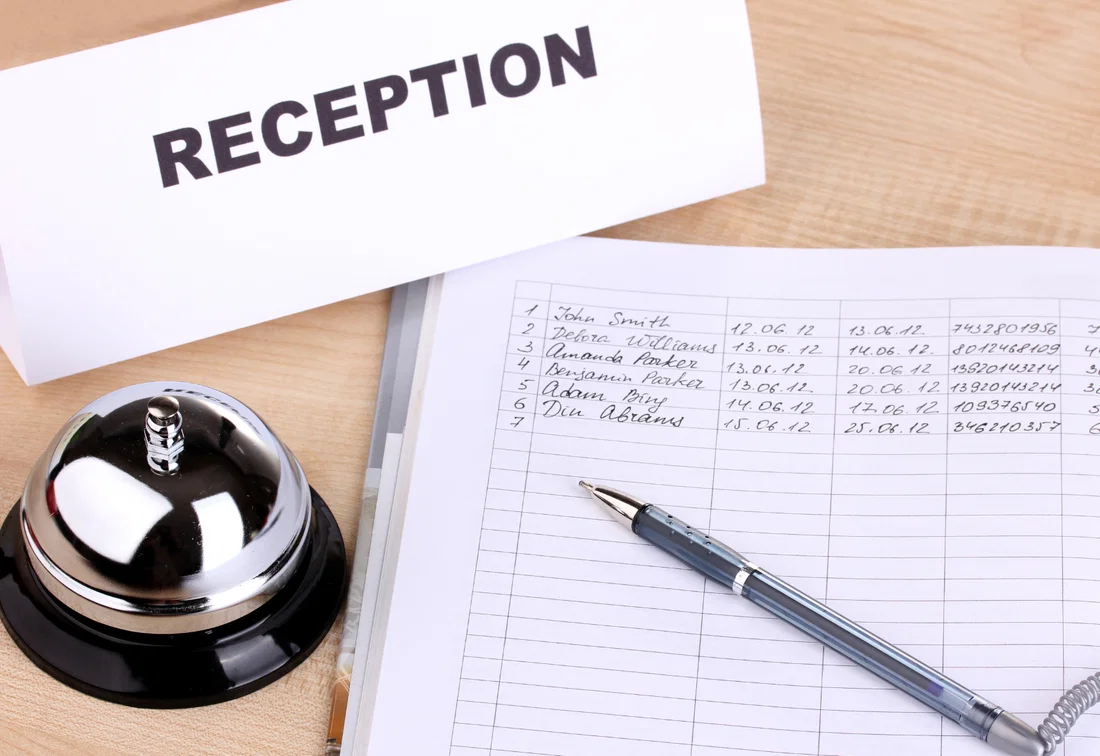 Balcão de recepção com placa RECEPTION, campainha e livro de visitas aberto com caneta para registo de visitantes.