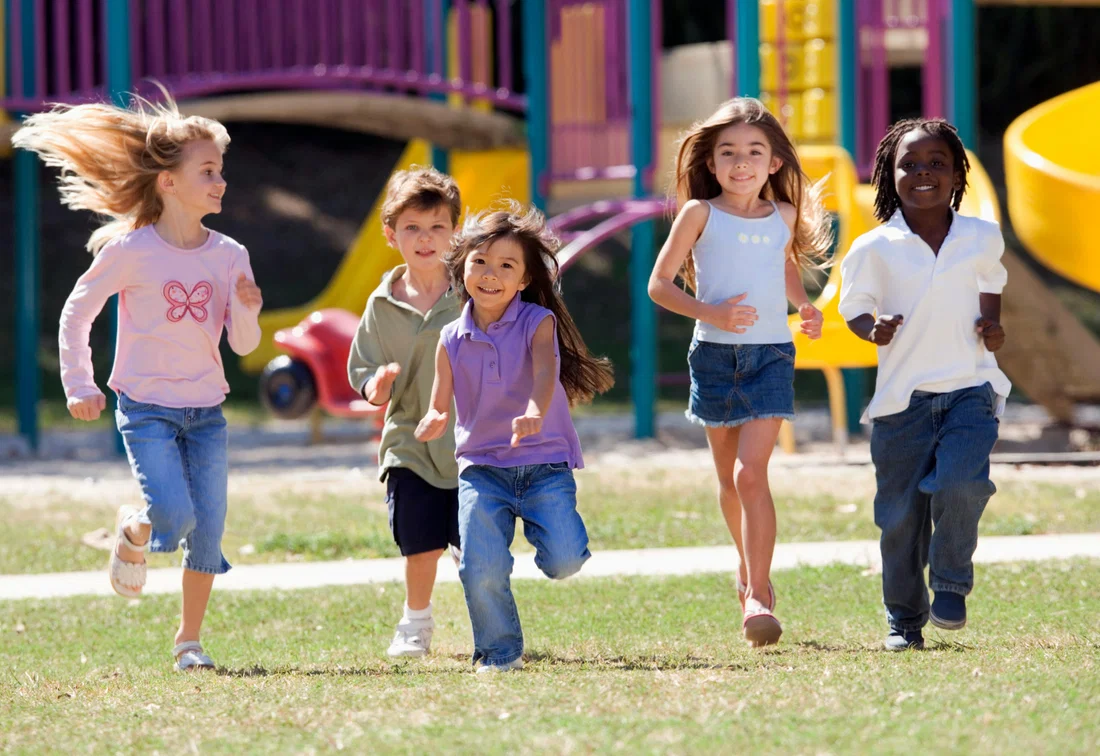 Niños con atuendos coloridos pasean por el patio de su escuela con juegos infantiles al fondo.