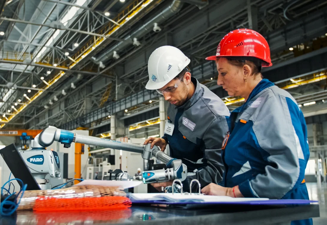 Dos operarios con cascos de seguridad realizan un control de calidad utilizando un brazo de medición 3D en una nave de producción.