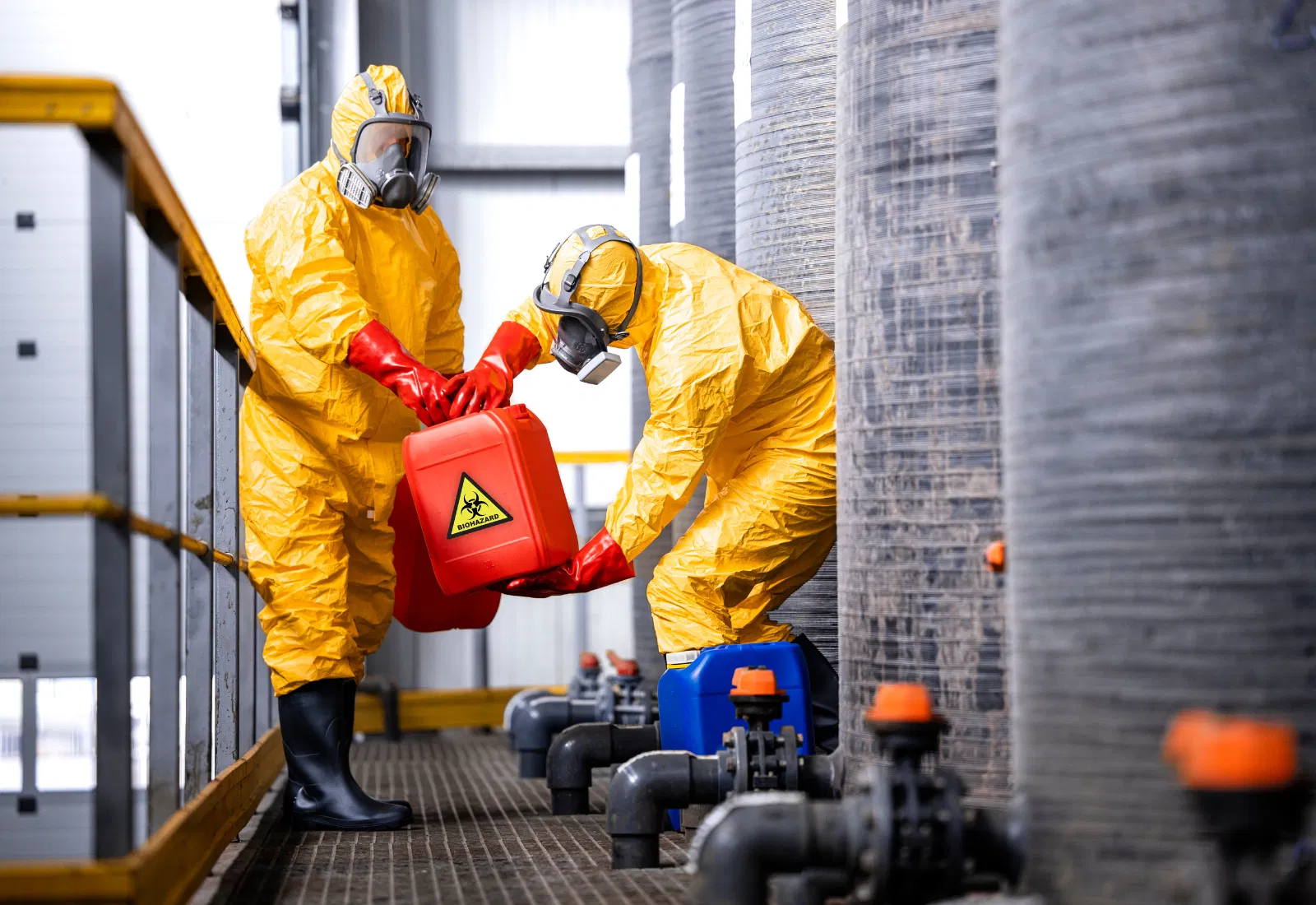 Dois funcionários vestindo fatos de proteção química amarelos com máscaras respiratórias movem um jerrycan vermelho com a etiqueta «perigo biológico» entre tanques de armazenamento e tubos numa fábrica.