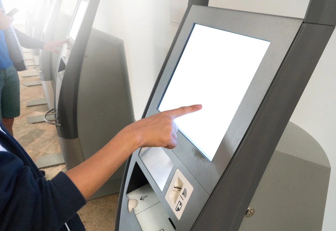 A hand using a self-service kiosk for check-in with touchscreen, NFC reader, and badge printer.