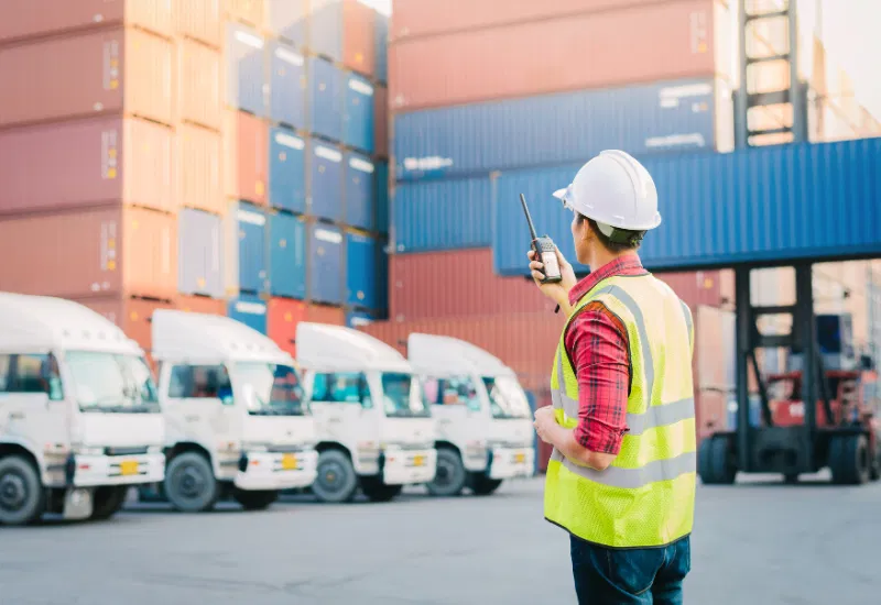 Arbeiter in einer Warnweste und einem Helm benutzt ein Funkgerät in einem Containerterminal, mit gestapelten Containern und geparkten Lastwagen im Hintergrund.