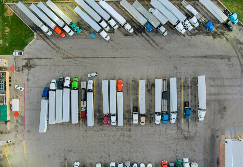 Eine Luftaufnahme eines LKW-Hofs mit mehreren geparkten Lastwagen und Anhängern, die in Reihen auf einem großen asphaltierten Gelände angeordnet sind.